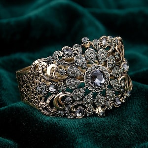 Puede incluir: Una pulsera de brazalete ancha con un intrincado diseño de filigrana metálica en tono dorado antiguo. La superficie está profusamente decorada con numerosos cristales pequeños de color gris oscuro y una piedra central más grande.