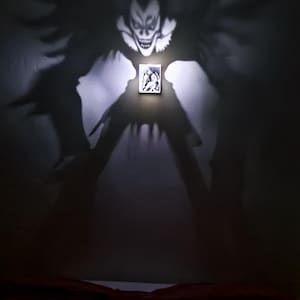 Op de afbeelding: Een kleine rechthoekige lichtbak met een zwart-witte grafische illustratie. Wanneer verlicht, projecteert het apparaat een grote, donkere en gedetailleerde schaduw van een mensachtig figuur met stekelig haar en een grijns op de muur.