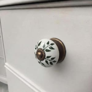 May include: A round white ceramic drawer knob featuring a green leaf pattern and a central bronze-toned metal accent. The knob is mounted on a white surface and includes a circular bronze-toned metal base plate that connects to the drawer front.