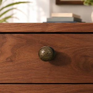 May include: A round, dark bronze-toned metal knob with a textured, mottled finish is centered on a horizontal wooden drawer front. The wood features a visible grain pattern in a medium brown tone, with a horizontal seam running above the drawer.