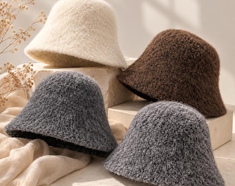 May include: Four bucket hats with a fuzzy, textured surface. One hat is cream-coloured, one is dark brown, one is charcoal grey, and one is a lighter grey. The hats are arranged in a group, showcasing their soft, fibrous material.