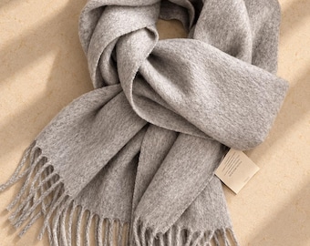 May include: A light grey scarf with a soft, textured finish and long fringe detailing at the ends. The scarf is loosely folded and features a small, rectangular white fabric tag attached to one side with printed text.