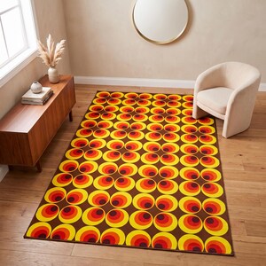 Op de afbeelding: Een rechthoekig vloerkleed met een herhalend geometrisch patroon van concentrische cirkels in tinten geel, oranje en rood tegen een donkerbruine achtergrond. Het ontwerp bestaat uit onderling verbonden cirkelvormige figuren.
