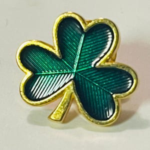 May include: A gold-toned shamrock pin with a green enamel finish. The three-leaf clover design has a gold border and stem. The pin is a classic symbol of luck and Irish heritage, suitable for St. Patrick's Day or everyday wear.