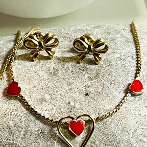 May include: A gold-toned necklace featuring three red heart charms and a matching pair of gold-toned bow-shaped stud earrings. The necklace chain is delicate and links the heart charms, with one larger heart frame containing a smaller red heart.