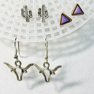 May include: A set of three pairs of earrings featuring a silver cactus, a silver bull's head, and a purple triangle.