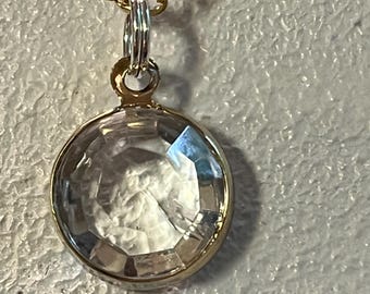 May include: A gold-toned necklace with a clear, faceted, round pendant. The pendant is set in a gold bezel and hangs from a delicate gold chain. The pendant's facets catch the light, creating a sparkling effect. The necklace is displayed against a textured, light-colored background.
