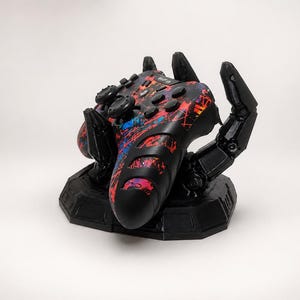 Può includere: Un controller per videogiochi nero con un vivace motivo a schizzi di vernice rossa e blu, sostenuto da un supporto a forma di mano robotica nera. Il controller presenta pulsanti standard, joystick e un pad direzionale su una base nera.