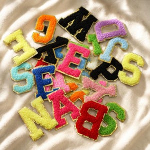 Puede incluir: Una colección de parches de letras texturizados y difusos en varios colores vibrantes, incluyendo amarillo neón, naranja, negro, rosa, azul, rojo y verde. Cada letra presenta un borde dorado y una superficie suave y afelpada para aplicar.