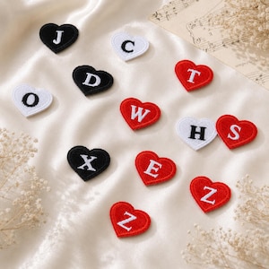 Puede incluir: Una colección de parches bordados en forma de corazón en negro, blanco y rojo. Cada parche presenta una letra mayúscula blanca o negra, incluyendo J, C, D, T, O, W, H, S, X, E y Z, dispuestos sobre una superficie de tela lisa y clara.