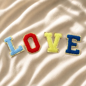 Peut inclure: Quatre lettres patch individuelles formant le mot LOVE. La lettre L est bleu clair, le O est rouge, le V est vert citron et le E est bleu foncé. Chaque lettre présente une surface texturée et duveteuse avec une fine bordure dorée.