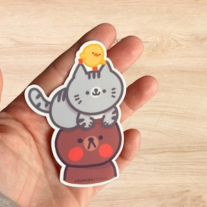 May include: A sticker featuring a brown bear with red cheeks, a gray cat with a yellow chick on its head, and the text "@lumiaustudio".