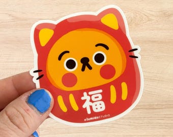 May include: A die-cut sticker featuring a Daruma doll design with a cat-like appearance. The sticker has a red and yellow colour scheme, with the Japanese character for "fortune" prominently displayed. The sticker is from @lumiastudio.