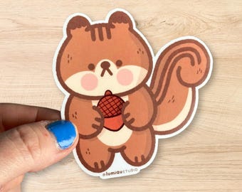 Puede incluir: Una ardilla de dibujos animados marrón y blanca sosteniendo una bellota marrón. La ardilla tiene una cola larga y tupida. La pegatina tiene el texto "@lumiqustudio" en la parte inferior.