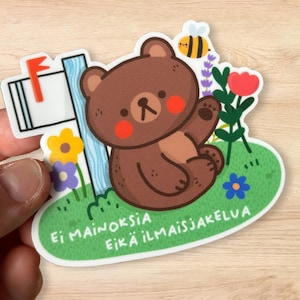 Peut inclure: Un autocollant représentant un ours brun de dessin animé avec des joues roses, saluant depuis une zone herbeuse avec des fleurs et une boîte aux lettres. Une abeille jaune et noire plane à proximité. Le texte "Ei mainoksia eikä ilmaisia" est imprimé.