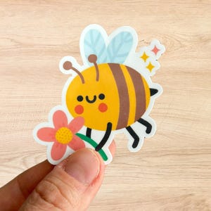 Könnte beinhalten: Ein farbenfroher Aufkleber mit einer Cartoon-Biene, die eine Blume hält. Die Biene ist gelb mit braunen Streifen, rosigen Wangen und blauen Flügeln. Die Blume ist rosa mit einem gelben Zentrum. Glitzerakzente sind enthalten.