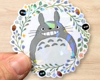 May include: A circular sticker featuring the character Totoro from the animated film, surrounded by a floral wreath. The sticker has a holographic, iridescent finish, and the character is grey with a white belly and a large smile.