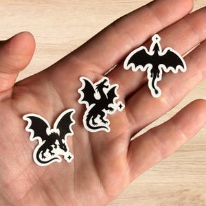 Könnte beinhalten: Drei schwarze Drachenaufkleber mit weißen Rändern werden gezeigt. Jeder Drache hat detaillierte Flügel und eine einzigartige Pose. Ein Drache steht aufrecht, ein anderer ist eingerollt und der dritte befindet sich in einer Flugposition. Ein kleiner Stern akzentuiert jeden Aufkleber.