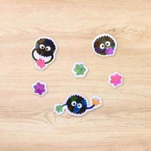 May include: A set of seven stickers featuring black and white fuzzy creatures with large eyes and colorful flowers. The stickers are on a light brown wooden surface.
