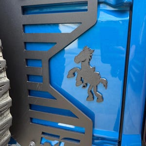 May include: A matte black metal automotive accessory panel mounted on a bright blue vehicle body. The panel features a cutout design with horizontal slats, a stylized rearing horse silhouette, and the letters RC embossed at the bottom corner.