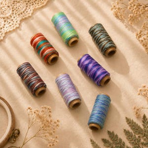 May include: Seven spools of embroidery thread arranged in a cluster. The spools feature variegated color combinations including green and blue, red and teal, dark brown and gold, purple, light lavender, and blue with silver metallic accents.