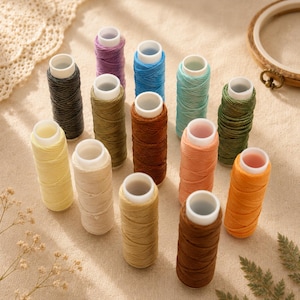 Può includere: Una collezione di quattordici rocchetti di filo in vari colori, tra cui giallo, beige, marrone chiaro, marrone, arancione, pesca, oliva, verde scuro, verde acqua, blu, viola e grigio antracite, ciascuno con un nucleo in plastica bianca.