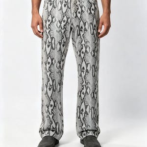 Könnte beinhalten: Lange Hose mit grau-schwarzem Schlangenledermuster. Die Hose verfügt über einen Standardbund mit Knopfverschluss, Gürtelschlaufen und ein klassisches Fünf-Taschen-Design. Der Stoff zeigt durchgehend einen detaillierten Reptilienschuppen-Druck.