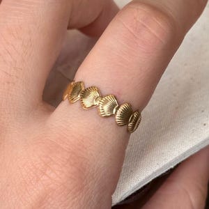 May include: A gold-toned ring featuring a repeating pattern of small, textured seashell shapes encircling the band. The text "Comfortable Fit" is printed in black above the ring, which is worn on a finger.