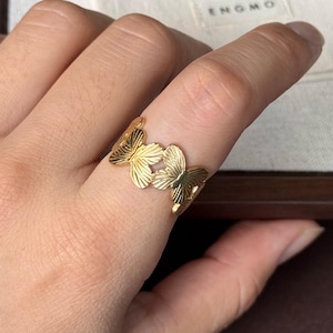 May include: A gold-toned ring featuring a series of connected butterfly shapes with textured wing details. The ring is worn on a finger, and the text Comfortable Fit and ENGMO are visible on a small fabric pouch in the background.