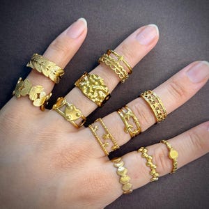 May include: A collection of ten gold-toned rings worn on fingers. The rings feature various designs including leaves, butterflies, hearts, stars, moons, scalloped edges, and textured surfaces. Each ring is crafted from a metallic material.