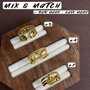 May include: Three gold-toned open rings displayed on a wooden holder. The top ring features a star cutout pattern, the middle ring has a beaded edge with star accents, and the bottom ring has a multi-band design with star motifs. Text reads MIX & MATCH &ndash; BUY MORE &middot; SAVE MORE, #5, #4, and #7.