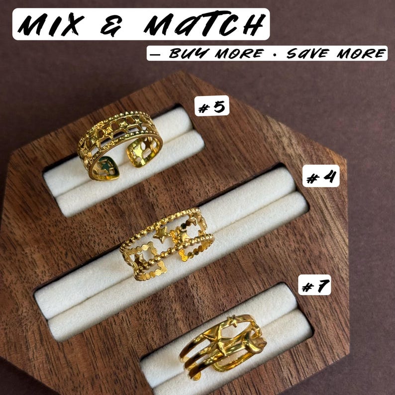 May include: Three gold-toned open rings displayed on a wooden holder. The top ring features a star cutout pattern, the middle ring has a beaded edge with star accents, and the bottom ring has a multi-band design with star motifs. Text reads MIX & MATCH &ndash; BUY MORE &middot; SAVE MORE, #5, #4, and #7.