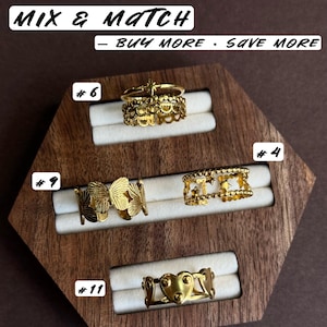 May include: Four gold-toned rings displayed on a wooden holder. Ring #6 features a star and scalloped band. Ring #9 has a butterfly motif. Ring #4 displays a star pattern. Ring #11 shows a heart design. Text reads MIX & MATCH - BUY MORE - SAVE MORE.