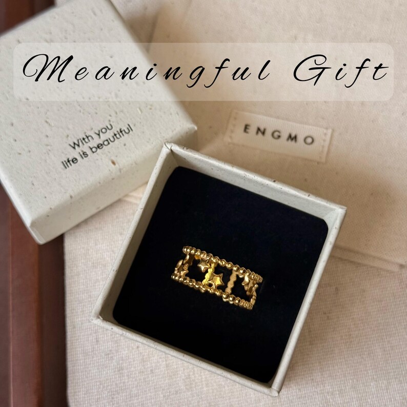 May include: A gold-toned ring featuring a cutout star pattern and beaded edges, presented in an open square box. The box lid is printed with the text Meaningful Gift and With you life is beautiful, while the box interior features the brand name ENGMO.