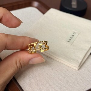 May include: A gold-toned ring featuring a double-band design connected by small star-shaped cutouts and beaded edges. The ring has an openwork structure with a polished finish, showcasing a celestial-inspired pattern throughout the band.