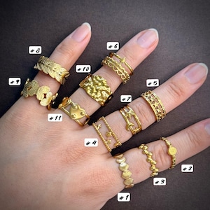 May include: A collection of eleven gold-toned metal rings worn on fingers, labelled with numbers 1 through 11. The rings feature various designs including leaves, butterflies, hearts, stars, moons, scalloped edges, and textured surfaces, all displayed together.