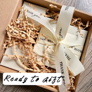 May include: A brown cardboard gift box filled with light brown shredded paper filler. Inside are white rectangular cards tied with a cream-coloured grosgrain ribbon printed with the words THANK YOU in black. The text Ready to Gift is visible.