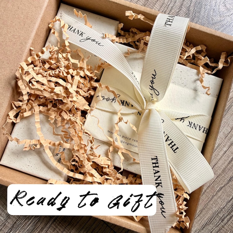 May include: A brown cardboard gift box filled with light brown shredded paper filler. Inside are white rectangular cards tied with a cream-coloured grosgrain ribbon printed with the words THANK YOU in black. The text Ready to Gift is visible.