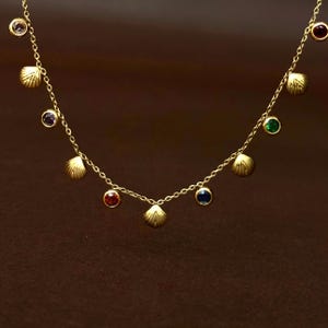 May include: A gold chain necklace featuring alternating charms of small, round, multi-coloured gemstones and textured gold seashell shapes. The gemstones include clear, purple, red, blue, green, and dark red stones set in gold bezels.
