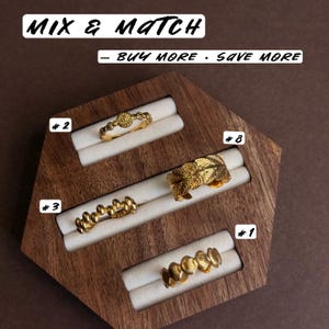 May include: Four gold-toned rings displayed on a hexagonal wooden stand with white velvet slots. The rings are numbered 1, 2, 3, and 8. Text at the top reads MIX & MATCH and BUY MORE &middot; SAVE MORE. The rings feature distinct textures including leaves, shells, and beads.