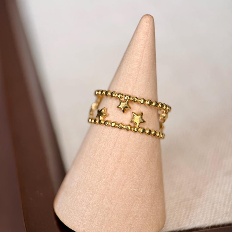 May include: A gold-toned ring featuring two parallel bands of small, uniform beads. Between the beaded bands, several small, five-pointed star shapes are evenly spaced, creating a delicate and open design that encircles the finger.