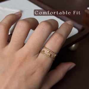 May include: A gold-toned ring featuring two parallel bands with a beaded texture, connected by small star-shaped accents. The ring is displayed with the text Comfortable Fit visible in the upper portion of the frame, indicating its design.
