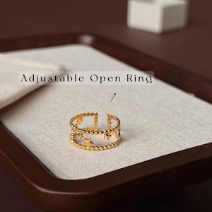 May include: A gold-toned adjustable open ring featuring two parallel bands connected by small star-shaped cutouts and a beaded texture along the edges. The text "Adjustable Open Ring" is printed above the jewellery piece with an arrow pointing down.