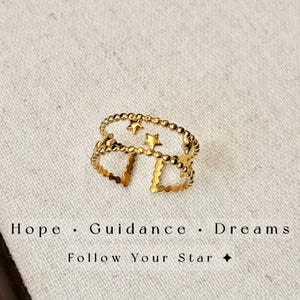 May include: A gold-toned open ring featuring two parallel bands connected by small star shapes. The ring has a beaded texture along the edges. Below the ring, the text reads: Hope &middot; Guidance &middot; Dreams. Follow Your Star.
