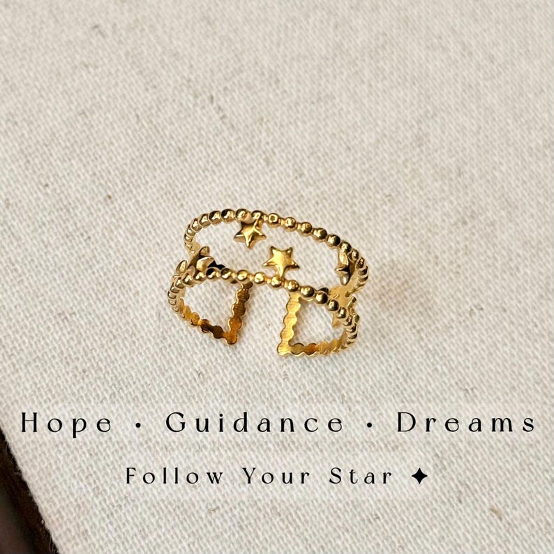 May include: A gold-toned open ring featuring two parallel bands connected by small star shapes. The ring has a beaded texture along the edges. Below the ring, the text reads: Hope &middot; Guidance &middot; Dreams. Follow Your Star.