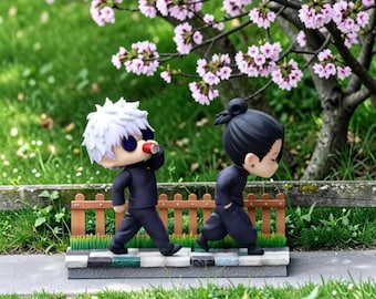 Puede incluir: Dos figuras de anime estilo chibi sobre una base de piedra frente a una pequeña valla de madera. Una figura tiene el pelo blanco puntiagudo y gafas oscuras, mientras que la otra tiene el pelo negro recogido en un moño. Ambas visten uniformes azul oscuro.