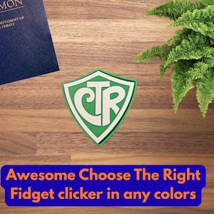 May include: A shield-shaped green and white fidget clicker featuring the letters CTR in the center. The object is displayed alongside a blue book and a green plant. The text at the bottom reads: Awesome Choose The Right Fidget clicker in any colors.