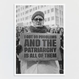 Könnte beinhalten: Ein Schwarz-Weiß-Foto einer Person mit Hut und Brille, die ein rechteckiges Schild mit dem Text hält: I GOT 99 PROBLEMS AND THE PATRIARCHY IS ALL OF THEM. Die Person ist von einer Menschenmenge vor einem großen Gebäude umgeben.