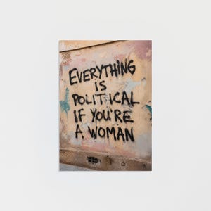 May include: A rectangular wall art print featuring a weathered, light-colored wall with peeling paint and a small vent at the bottom. The black spray-painted text reads: Everything is political if you're a woman. The surface has a textured, aged appearance.