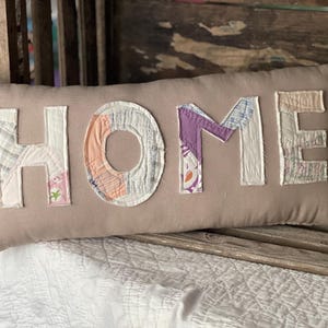 May include: A rectangular beige pillow with the word "HOME" appliquéd in patchwork fabric. The letters are in various colors and patterns, including white, purple, and peach. The pillow is resting on a wooden surface.
